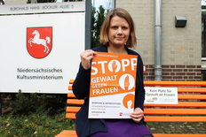Kultusministerin Hamburg auf der orangefarbenen Bank mit einem orangefarbenen Plakat, welches auf bestehende Hilfangebote hinweist.