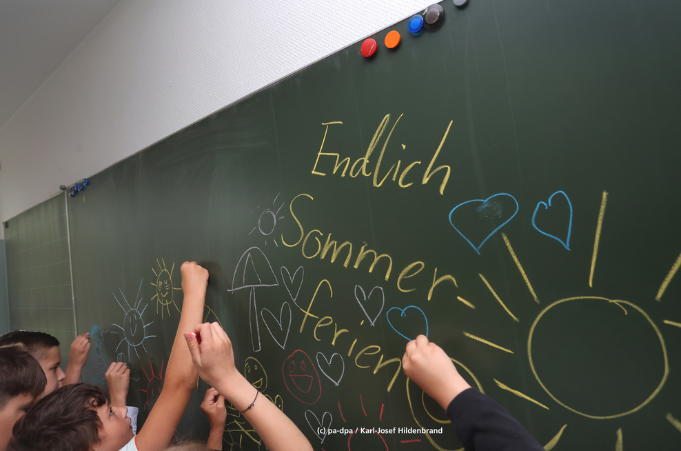 Kinder schreiben auf einer bunten Schultafel das Wort Sommerferien